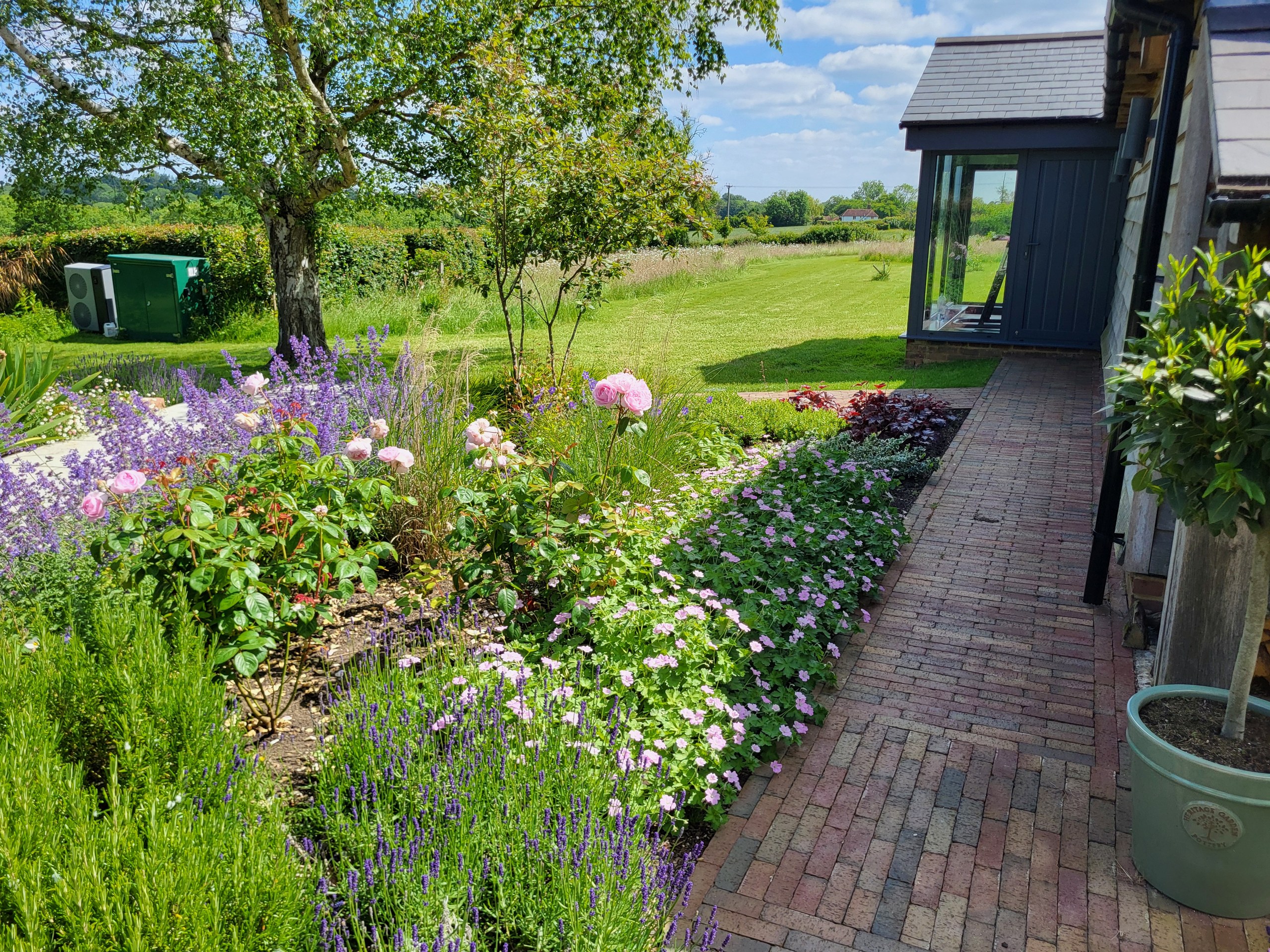 Brick path beside a house with a colourful flower border and lawn beyond in Benenden.