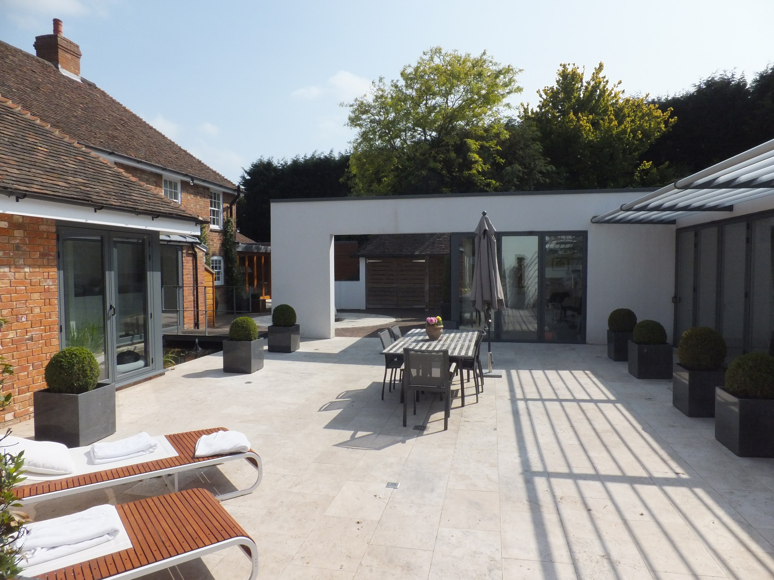 Paved courtyard with outdoor dining furniture, sun loungers and clipped shrubs beside a brick house and modern extension in Borden.