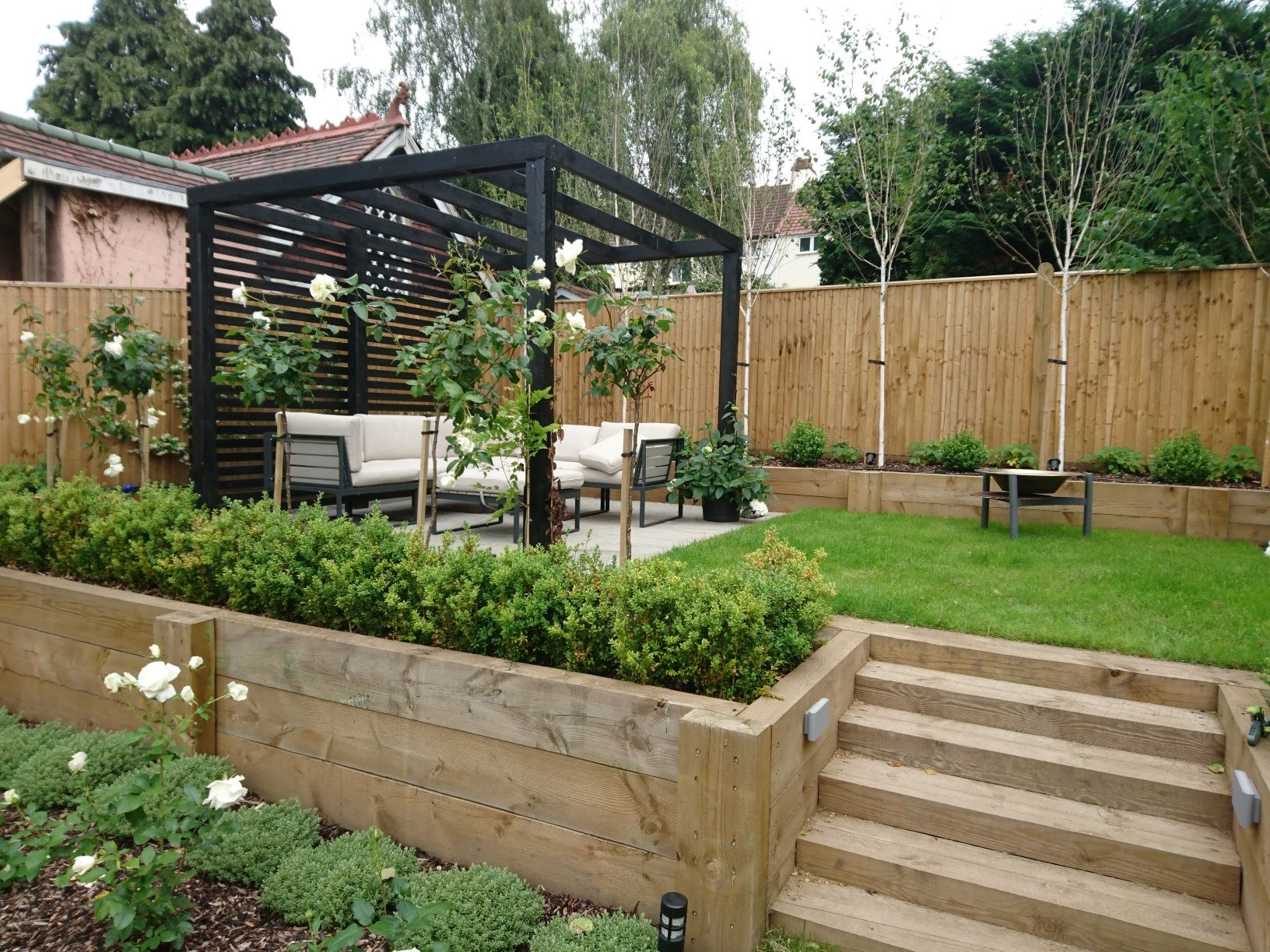 bromley-pergola-raised-lawn-overview-wealden-country-landcraft Black pergola with outdoor seating beside a raised lawn, stone steps and planted beds in Bromley.