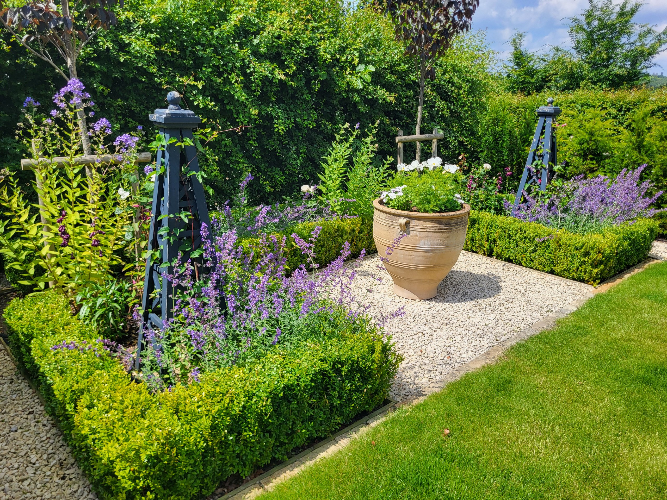 Blue plant obelisks and a large planted pot beside gravel paths and clipped border hedges in Goudhurst.
