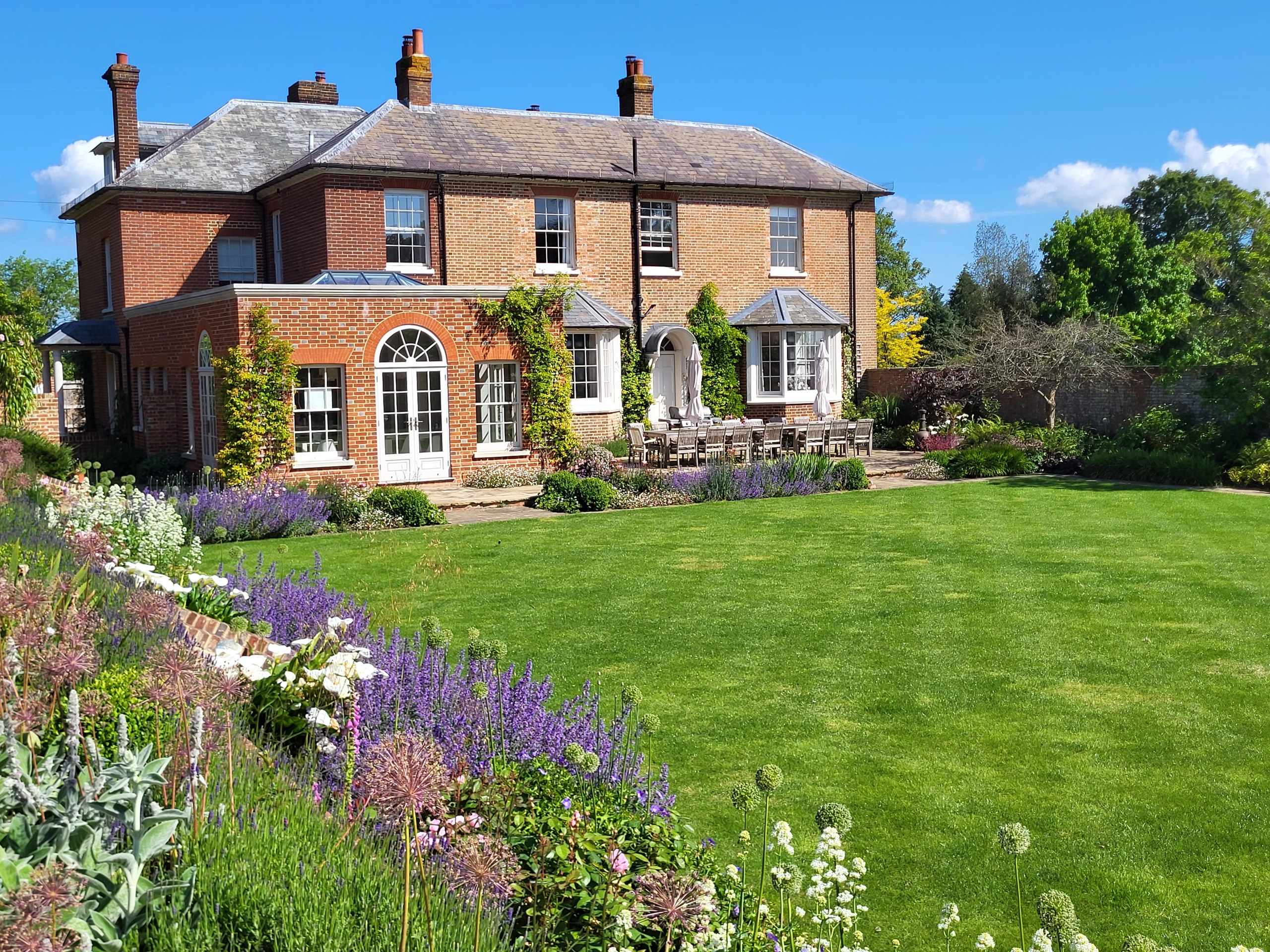 Broad lawn and flowering borders in front of a brick country house in Matfield.