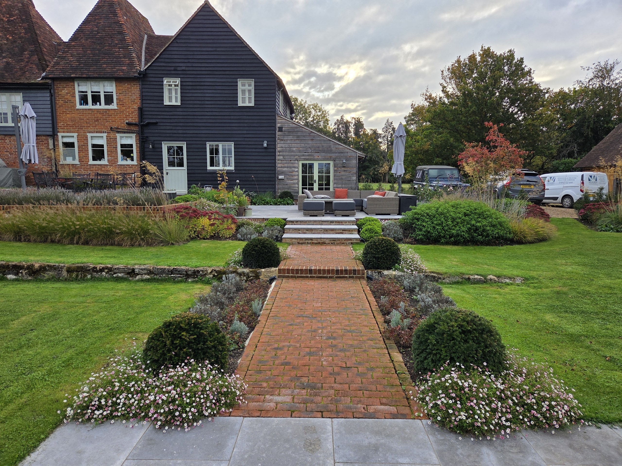 Straight brick path between clipped shrubs and flowering borders leading up steps towards the house in Sutton Valence.